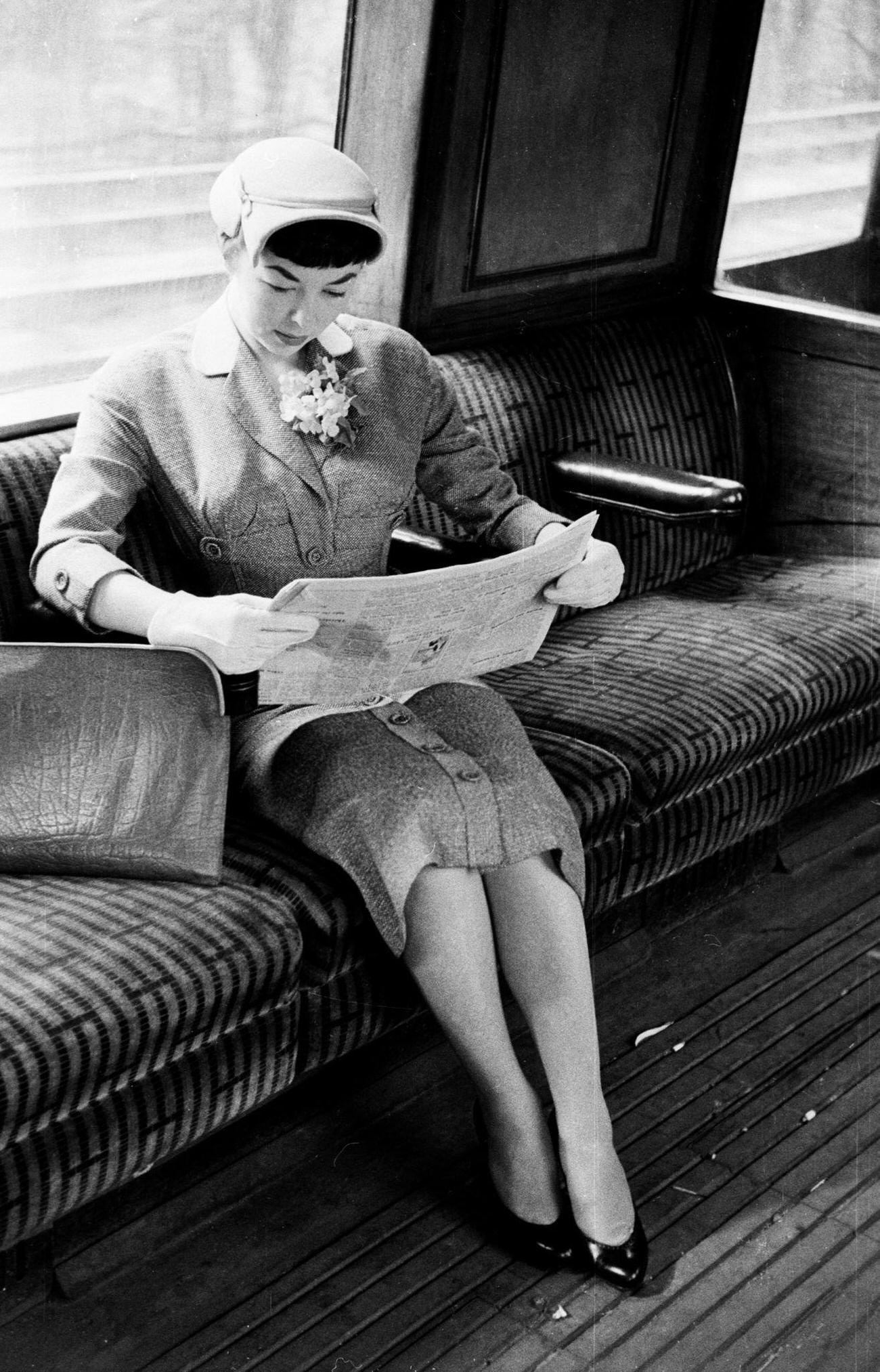 A woman wearing a grey tweed suit and reading a newspaper on the train journey to work, 1954