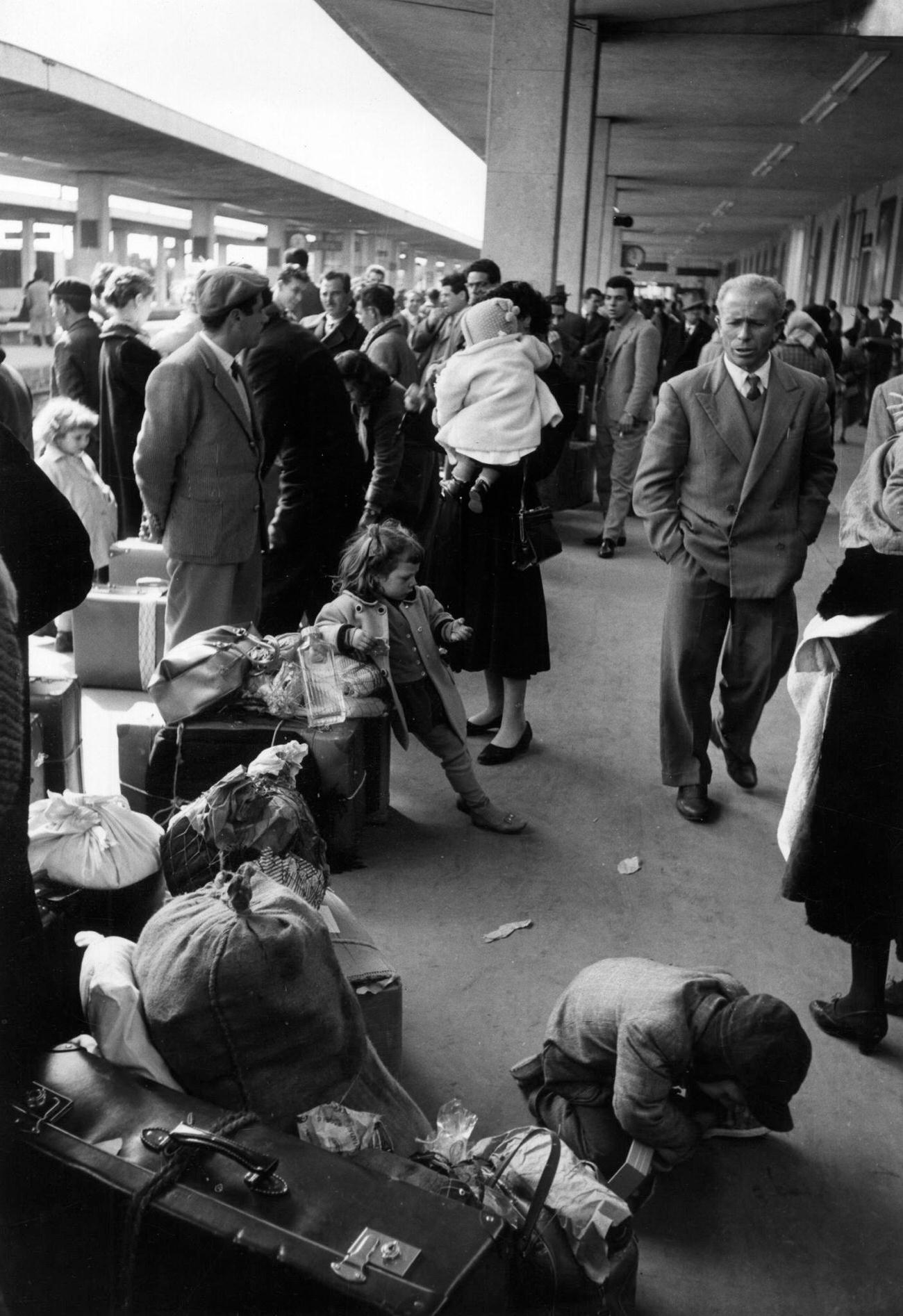 Families of emigrants with packs and luggage waiting for a train at the railway station. Catania, March 1958