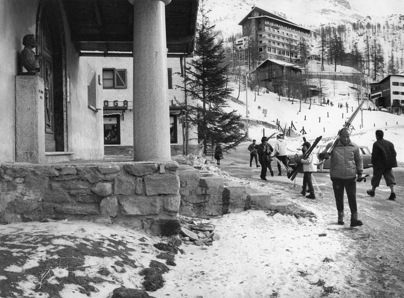 Some skiers, with the skis on their shoulders. walking in front of the new church founded by Don Giuseppe Vietto, the odd priest of Cervino. Valtournenche, 1964