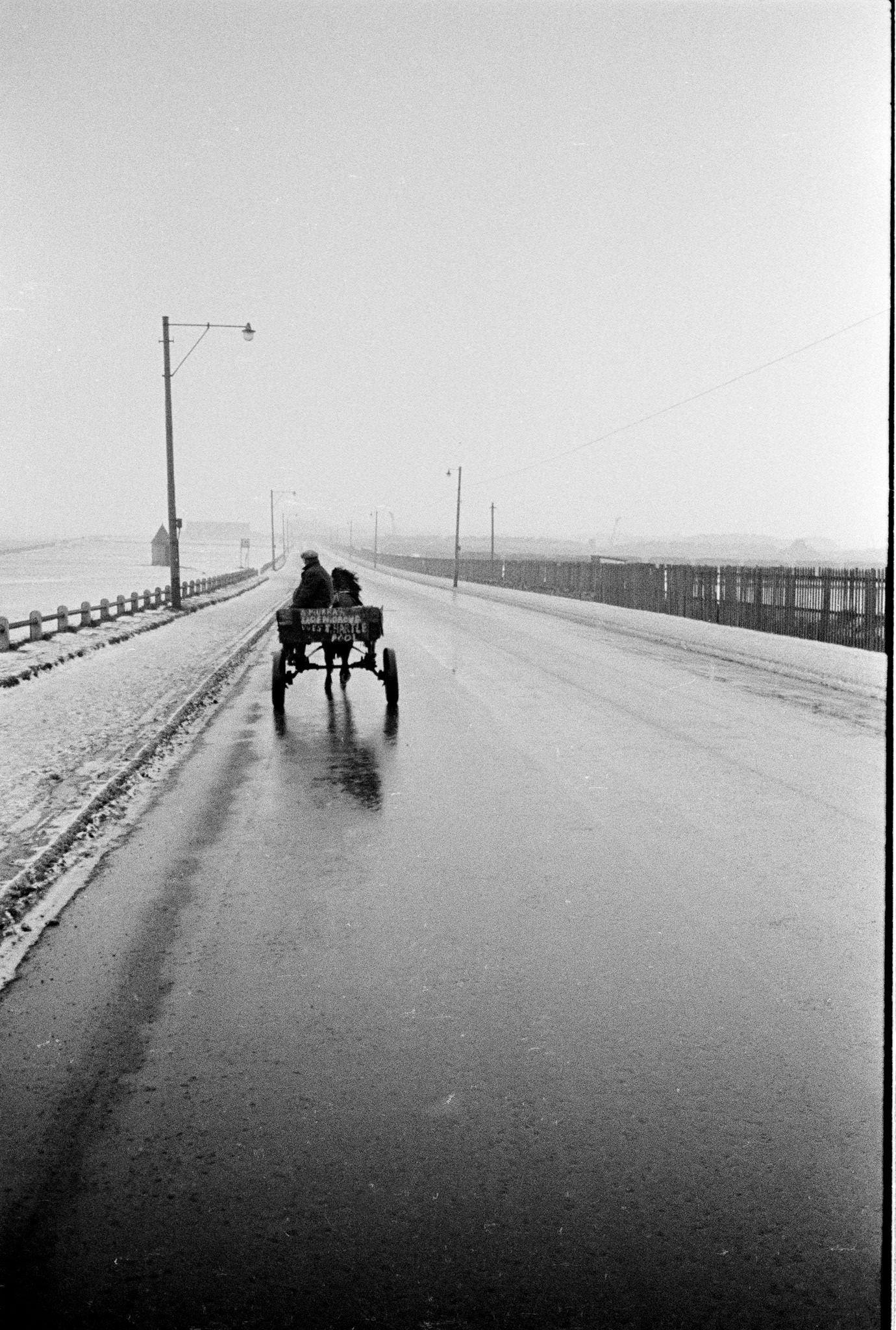 A man rides on a horse drawn cart along the coast road between Seaton Carew beach and Hartlepool in County Durham, northeast England during winter 1962.