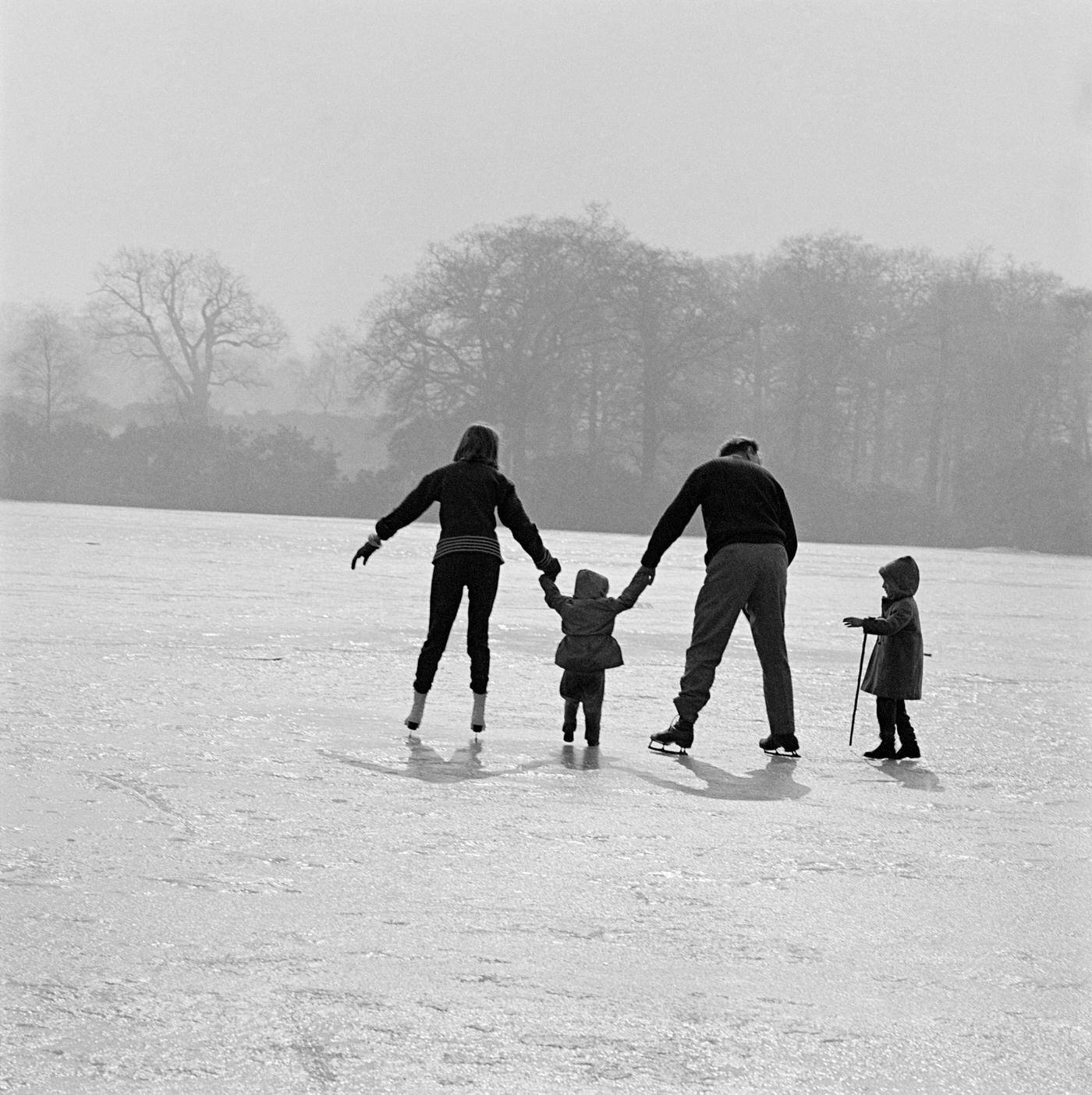 Skating in Richmond Park, London, 1962. A man and woman on skates helping two small children to walk on the ice on a pond or lake in Richmond Park in winter.