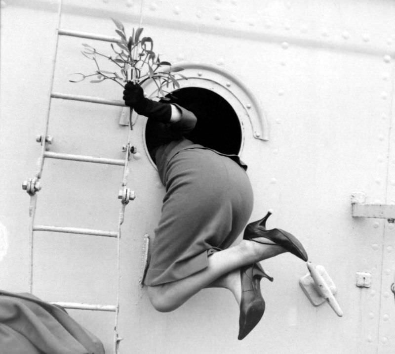 A woman, holding a mistletoe, got pulled through a porthole by her sailor husband for a homecoming kiss as the crew of HMS Bulworth returned for Christmas holidays, 1962. (Reg Lewis)