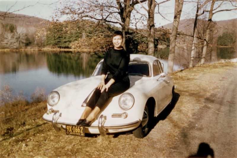 A stylish young lady posing on a Porsche 356 B Coupé by the side of a lake on a sunny autumn's day. The car is registered in New York State with 1960 licence plates, 1960