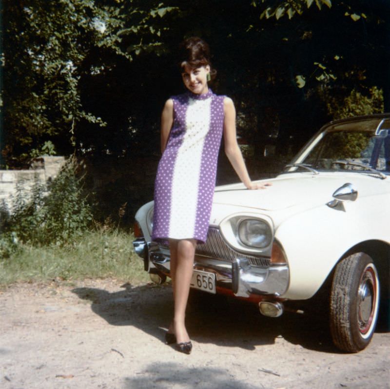 A brunette lady in a purple and white dress posing with a Ford Taunus 17M in summertime. The Taunus is equipped with amber fog lights and bullet-style wing mirrors, 1963