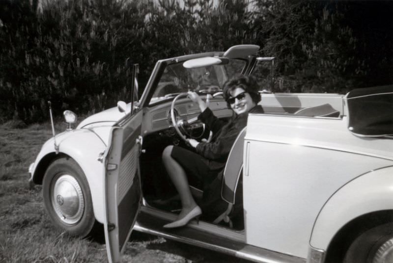 A cheerful lady posing in the driver's seat of a VW 1200 Cabriolet in the countryside. The car is equipped with fender guides and an additional wing mirror, 1963