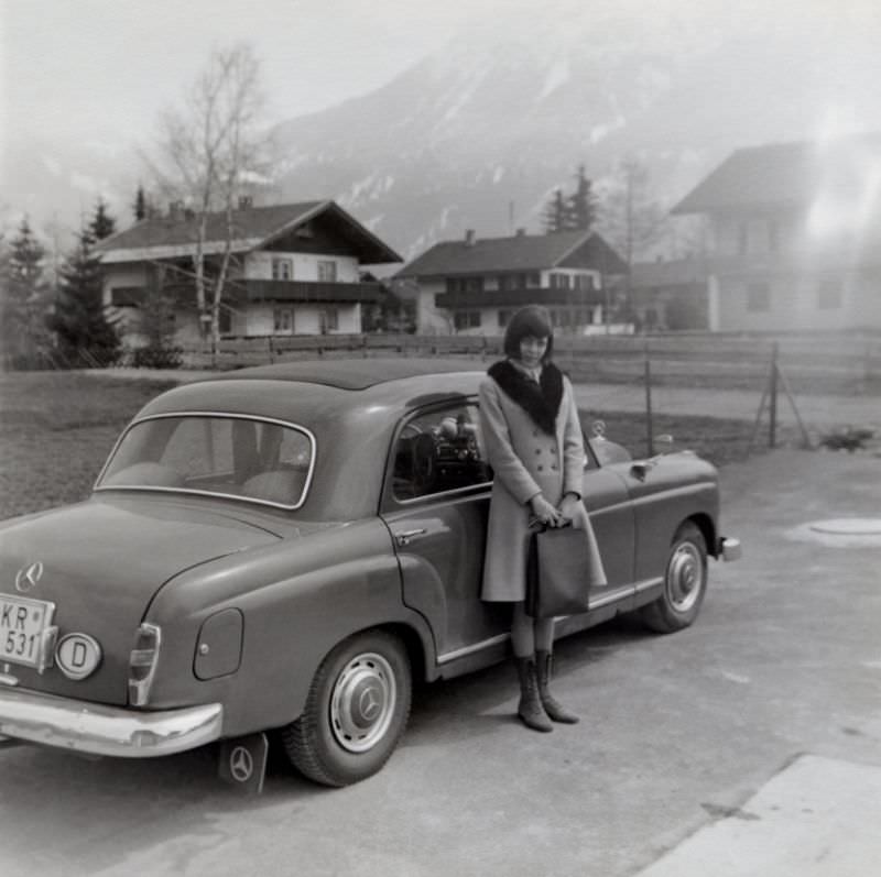 A young brunette lady posing with a Mercedes-Benz 180 in an Alpine setting. The car is equipped with a full-length folding canvas sunroof and registered in the West German town of Krefeld, 1963
