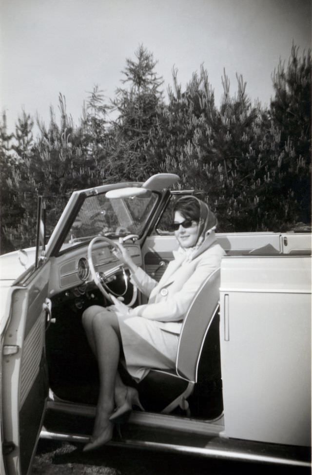 An elegant lady posing in the driver's seat of a VW 1200 Cabriolet in the countryside. The print is dated "1963" on reverse