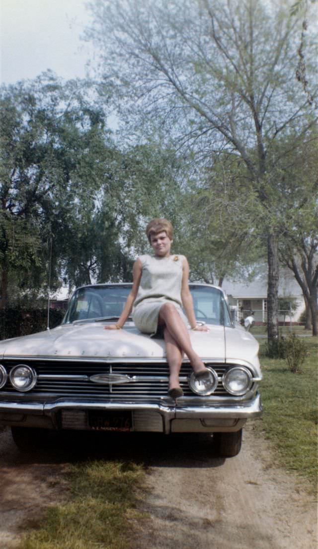 A young blonde lady in a sleeveless white dress posing on the bonnet of a 1960 Chevrolet Impala in the drive of a private home. The words "Nancy 1964" are handwritten on reverse