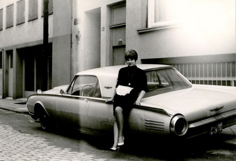 A brunette lady posing with a 1961 Ford Thunderbird in a cobbled downtown backstreet, 1965