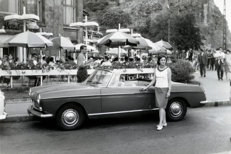 A brunette lady posing with a Peugeot 404 Cabriolet in front of a café in a busy city street, 1965