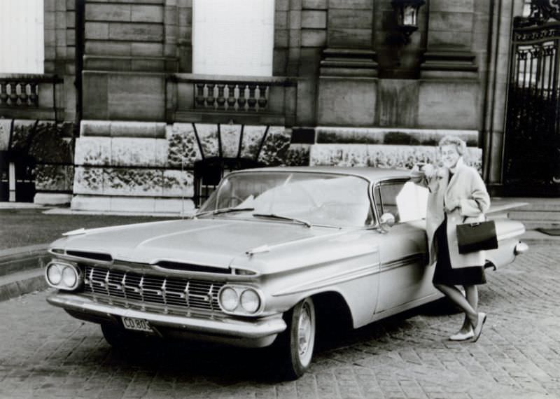 A cheerful blonde lady posing with a 1959 Chevrolet Impala Sport Coupe in front of a classical building, 1960