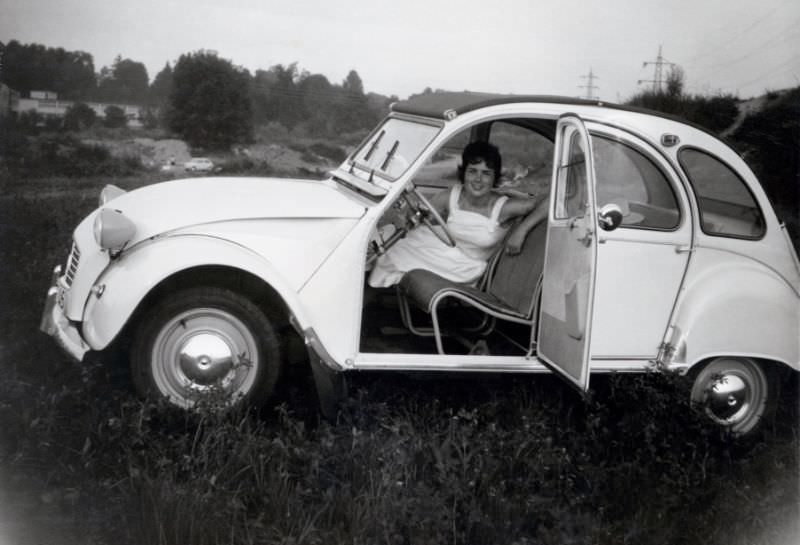A cheerful brunette lady in a white summer dress posing in the hammock-style seats of a Citroën 2CV, 1965