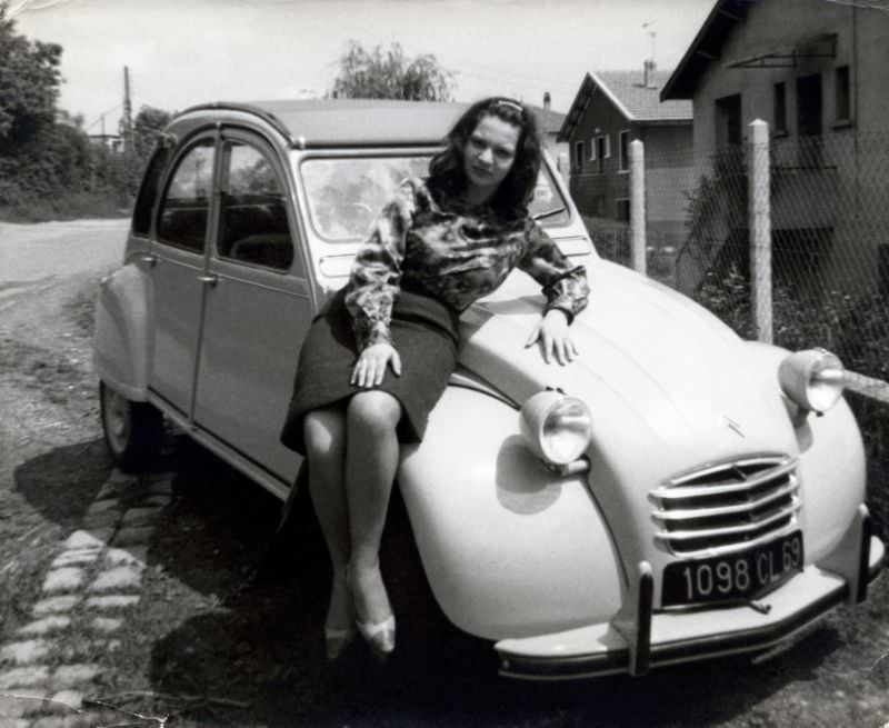 A cheerful brunette lady posing on the fender of a Citroën 2CV in a suburban street. The car is registered in the French département of Rhône, 1965