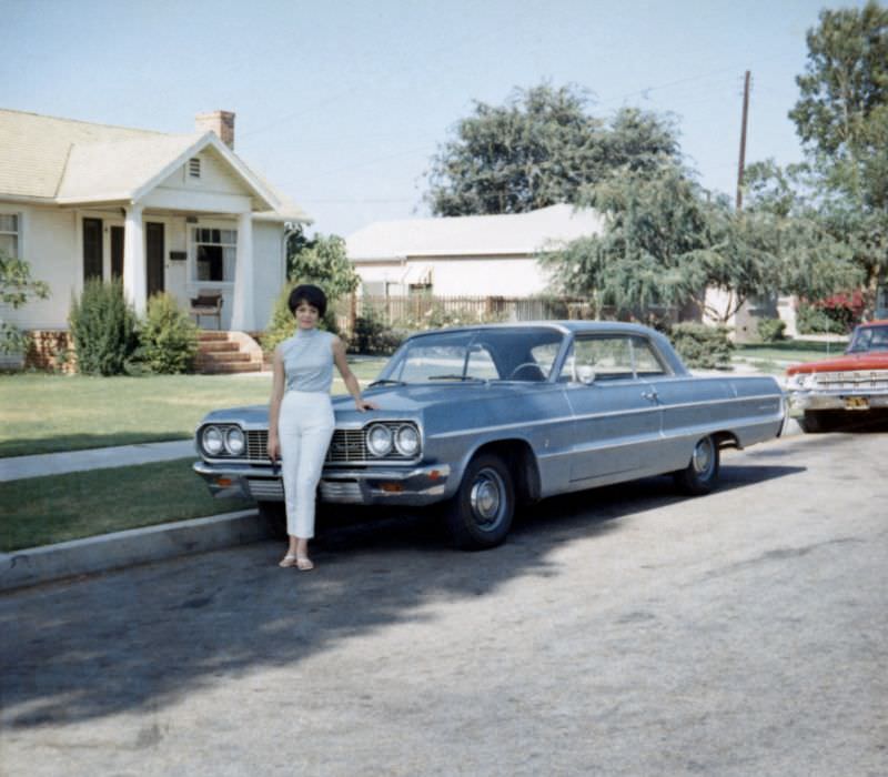 A young lady posing with a 1964 Chevrolet Impala Sport Coupe in a suburban street in summertime, 1965