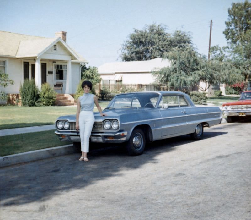 A young lady posing with a 1964 Chevrolet Impala Sport Coupe in a suburban street in summertime, 1965