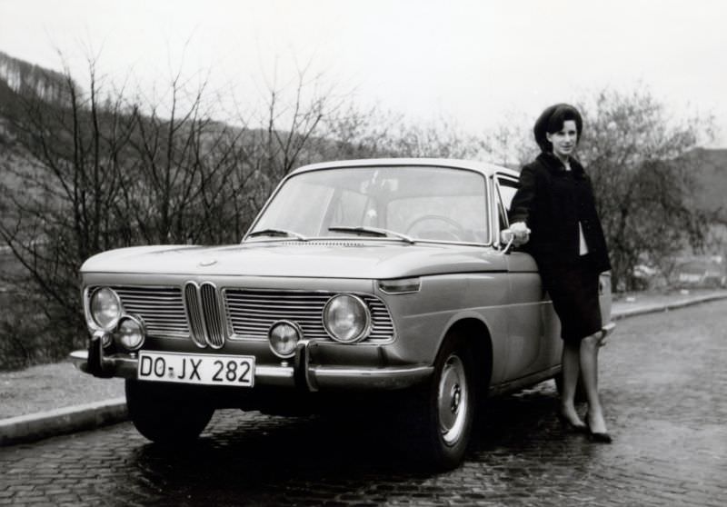 An elegant lady with a beehive hairdo posing with a BMW 1800 on a cobbled road in the countryside. The car is registered in the West German city of Dortmund, 1965