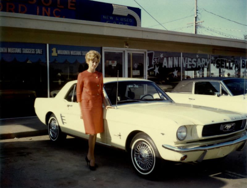 A blonde lady in a red female suit posing next to a 1966 Ford Mustang at a Ford dealership, 1966