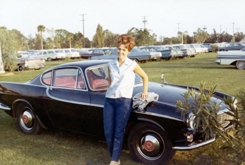 A cheerful young lady with a beehive hairdo posing with a maroon-colored Volvo P1800 in a parking lot. The words "Hannelore – Los Angeles 1966" are handwritten on reverse