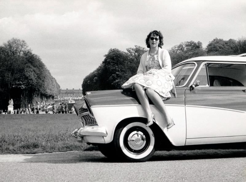 A dark-haired lady in a patterned dress posing on the bonnet of a Ford Taunus 17M De Luxe. The Palace of Versailles is visible in the background, Paris, April 1960