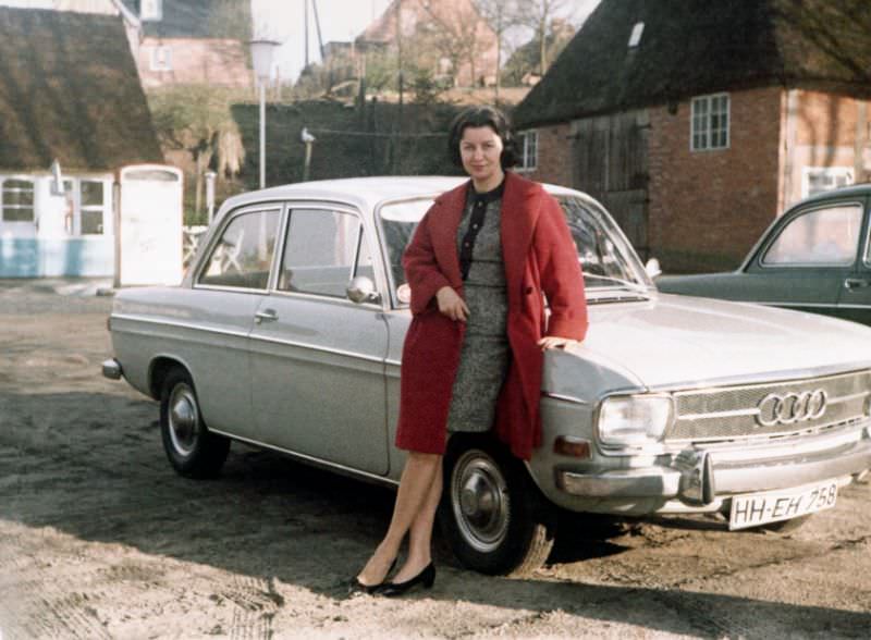 A brunette lady in a red coat posing with an Audi F103. The car is registered in the city of Hamburg, 1967