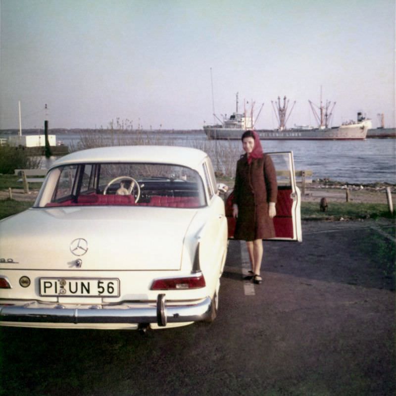 A lady wearing a brown coat and a red headscarf posing at the passenger-side door of a Mercedes-Benz 190. The car is registered in the West German district of Pinneberg, May 9, 1967