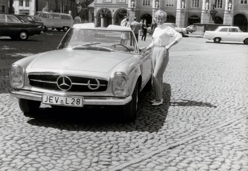 A blonde lady posing with a Mercedes-Benz Pagoda convertible in a cobbled old-town square. The car is registered in the West German town of Jever, 1968