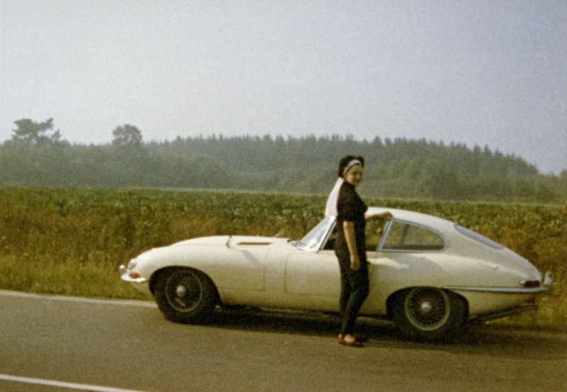 A fashionable lady posing with a Jaguar E-Type on the side of a country road in summertime, 1968