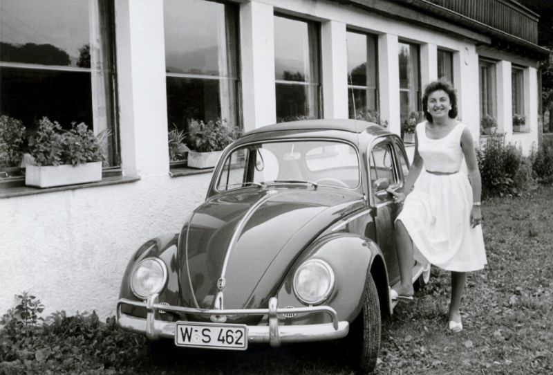 A cheerful brunette lady posing with a Volkswagen VW 1200 in front of a large building, possibly a hotel. The car is registered in the West German town of Wuppertal, 1962