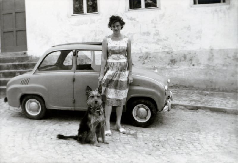 A cheerful brunette lady and her dog posing with a slightly shabby-looking Goggomobil T 250 in a cobbled street in summertime, 1960s