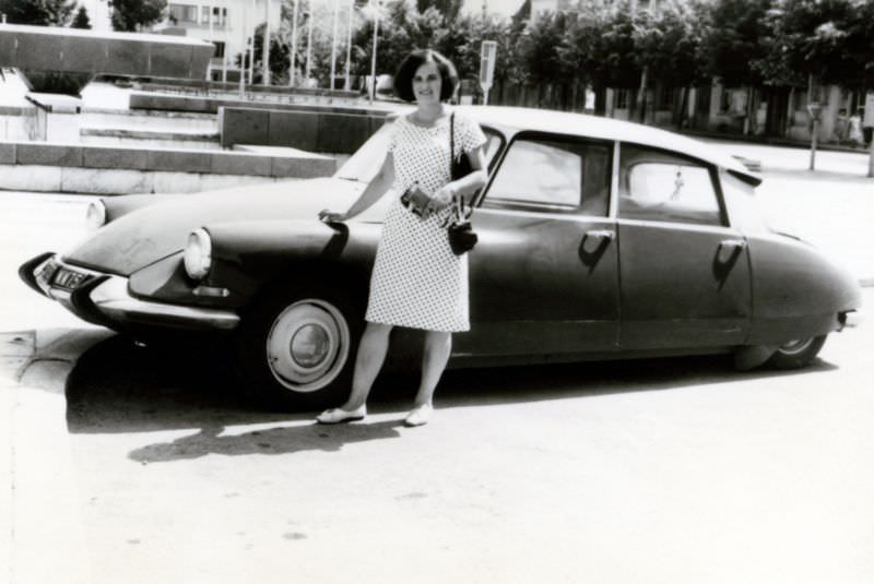 A cheerful lady in a polka dot dress posing with a Citroën in a city square in summertime, 1960s