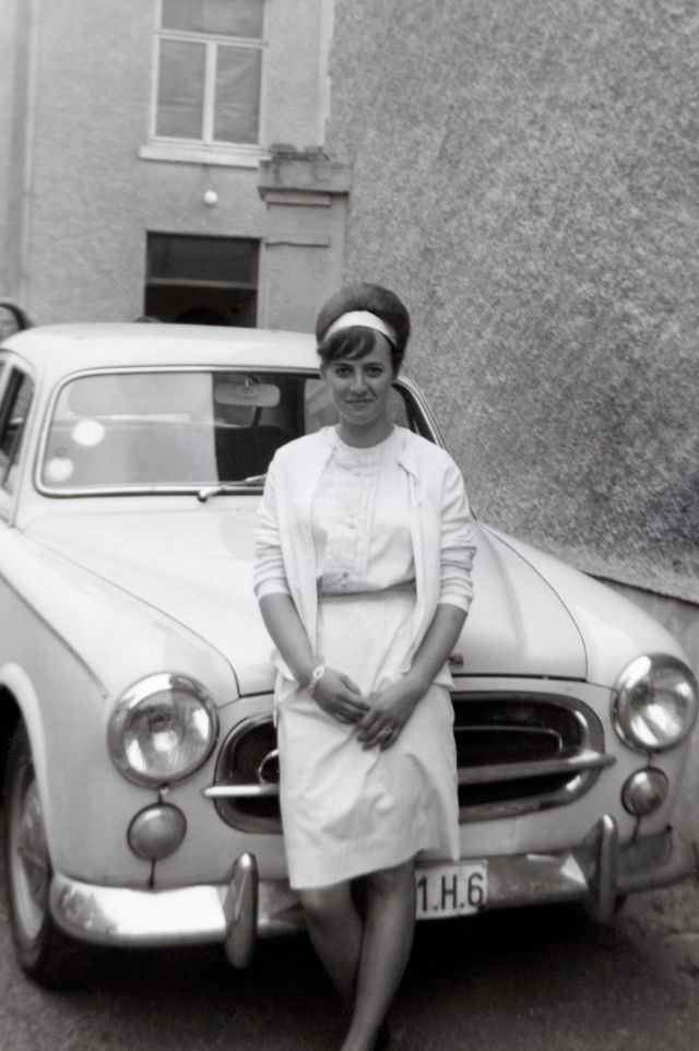 A cheerful lady with a beehive hairdo posing with a Peugeot 403 in an urban backyard. She is wearing a white skirt, blouse, cardigan and hairband. The car is registered with Belgian licence plates, 1960s
