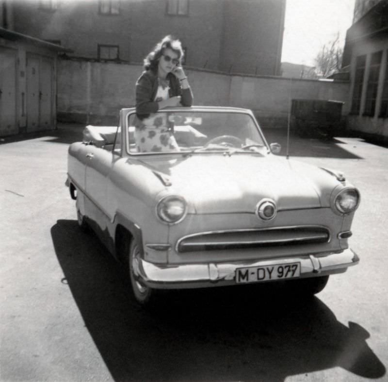 A young lady in a floral dress posing with a Ford Taunus 15M Cabriolet in summertime. The car is registered in the West German city of Munich, 1960s