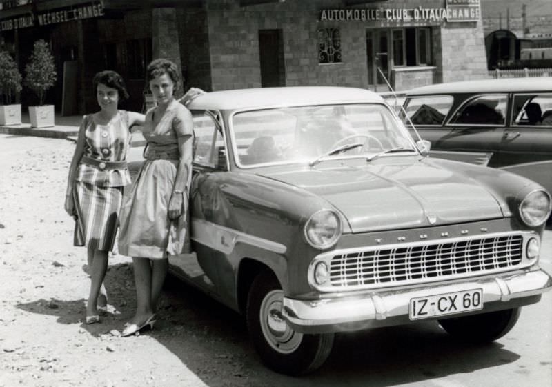 Two fashionable ladies posing with a Ford Taunus 12M in the parking lot of a railway station in summertime somewhere in Italy. The two-tone car is registered in the West German district of Itzehoe, 1960s