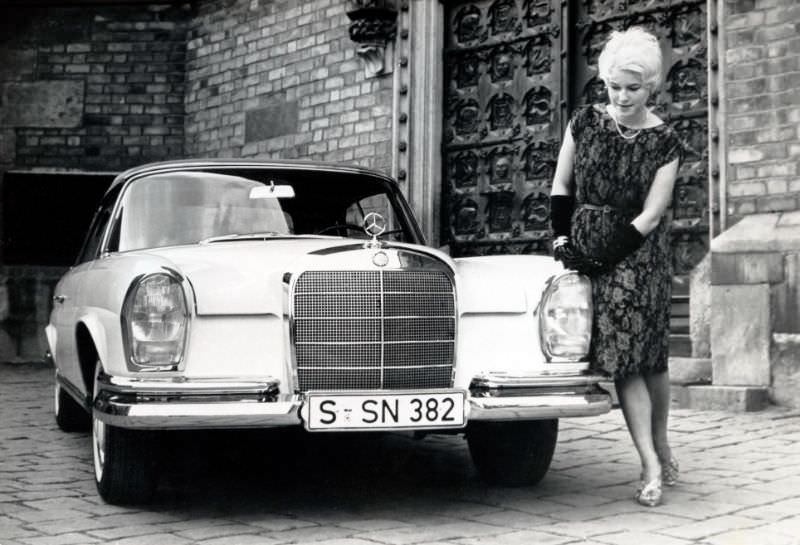 A platinum-blonde lady wearing a sleeveless floral dress and black velvet opera gloves posing with a Mercedes-Benz 220 SE Cabriolet in front of a medieval cathedral. The car is registered in the city of Stuttgart, 1962