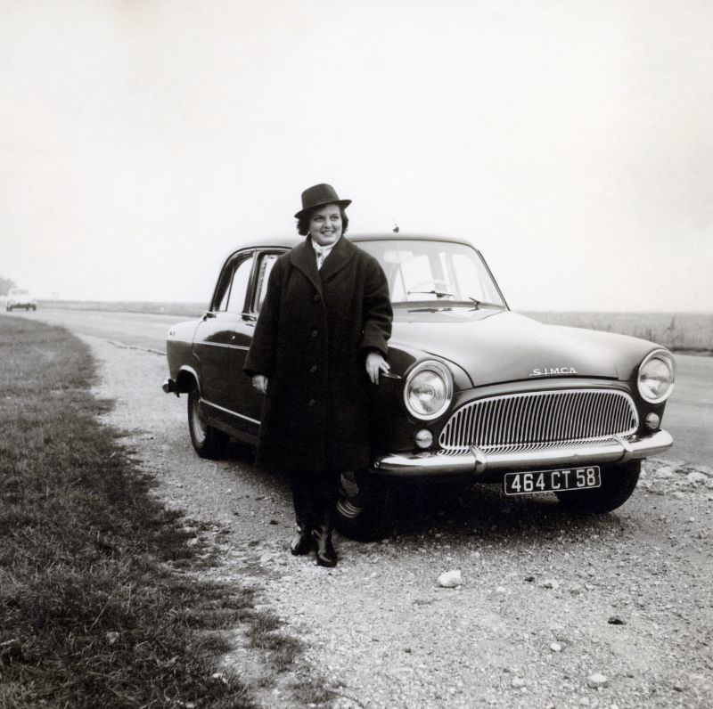 A stylish lady in a dark coat, hat, and boots posing with a Simca Aronde on the side of a country road. The car is registered in the French département of Nièvre, 1962