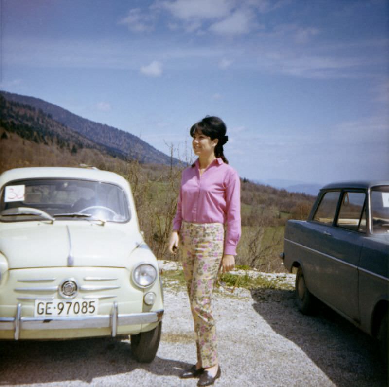 An elegant lady posing with a Fiat 600 in a graveled parking lot in the countryside. The car is registered in the city of Geneva, 1962