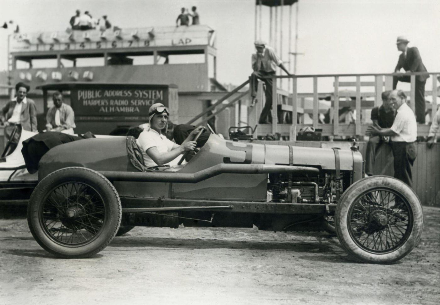 Joe Crocker poses in a race car at the speedway with a group of spectators milling about in the background, 1935.