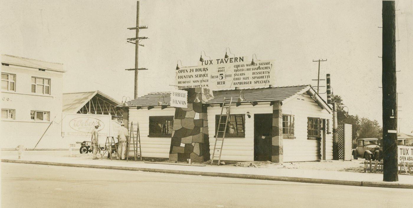 Exterior of Tux Tavern, a small building with a Tux Tavern sign on top, wood siding and a ladder leading against it, 1930s.