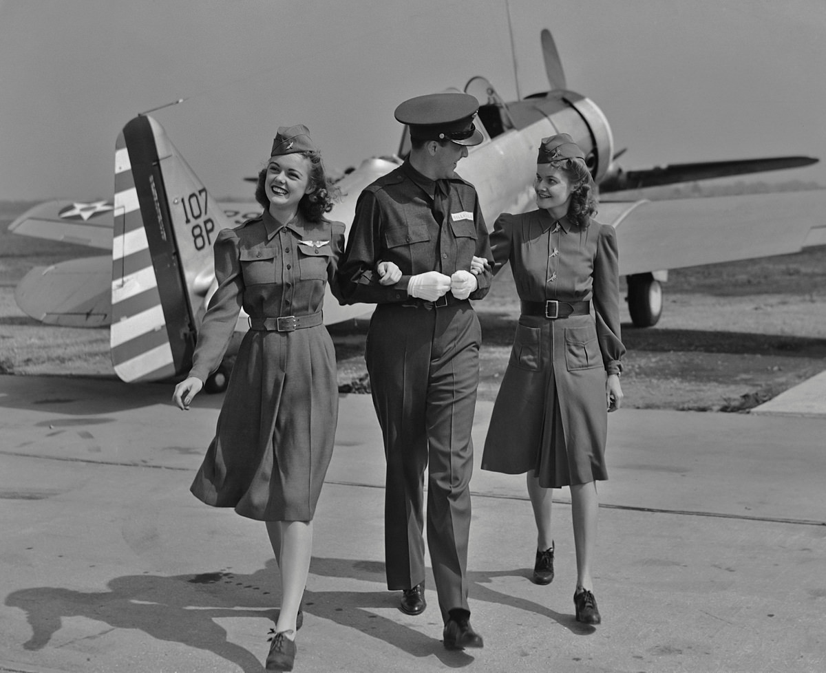 In the 1940s, a male flight attendant walks with his arms linked with two female flight attendants in front of a small plane.