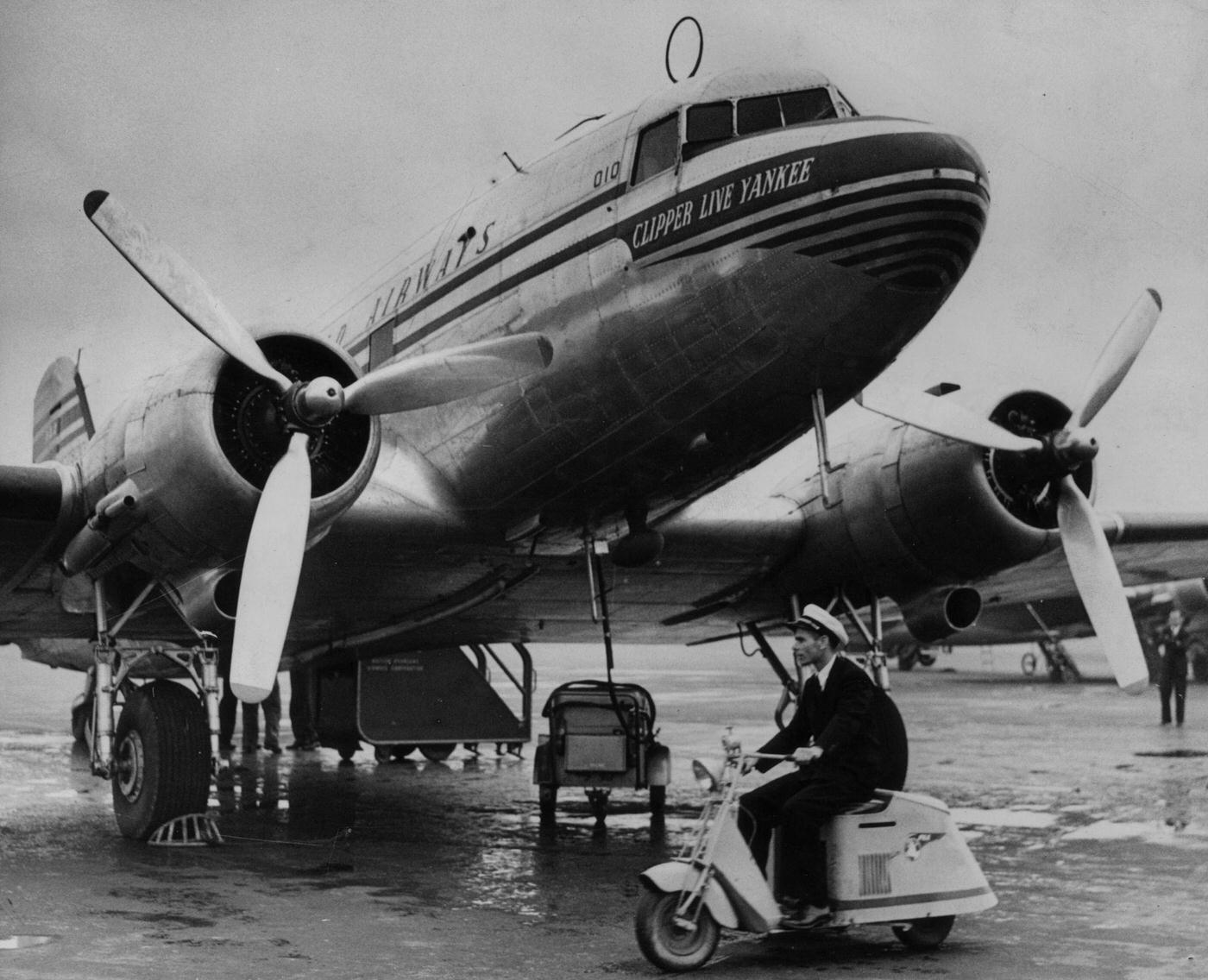 A motor-tricycle 'scooter' is being used for quick communication between airport buildings and planes at London Airport (Heathrow). One of the planes on the tarmac is a Pan-Am Clipper Line Yankee.
