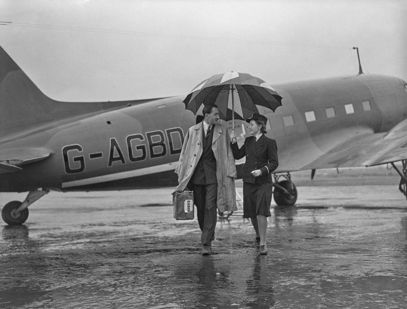 A female travel clerk with a large umbrella provides shelter for a passenger arriving during heavy rain on an incoming British Overseas Airways Corporation (BOAC) flight at an airport in England.