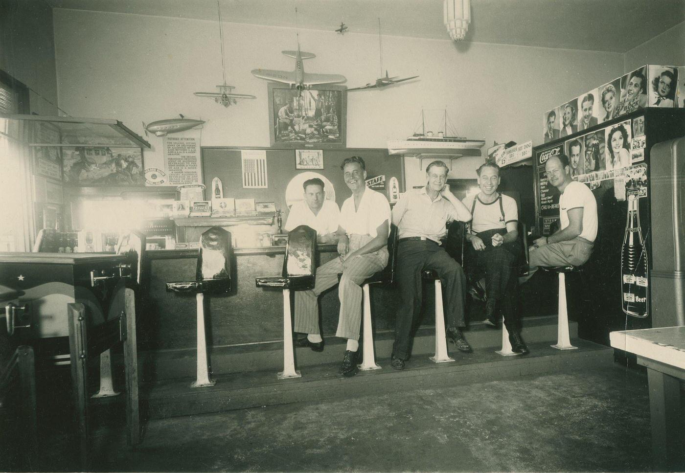 Four men are shown sitting on stools at a counter, with a man behind the counter.