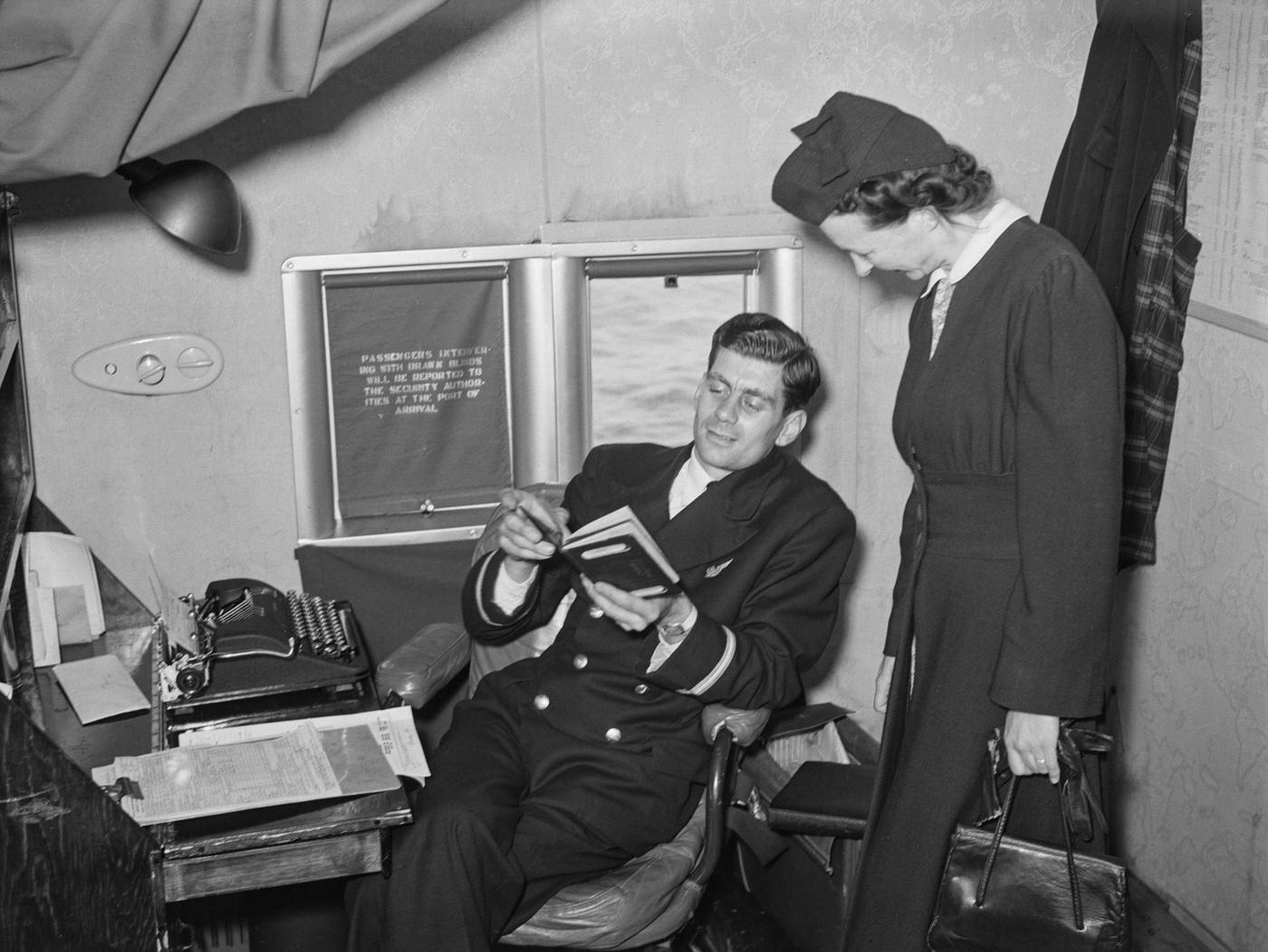 Purser K Everall is assisting a female passenger with a passport query aboard a British Overseas Airways Corporation (BOAC) flying boat flight from England during World War II on 31st August 1942, while aboard the plane.