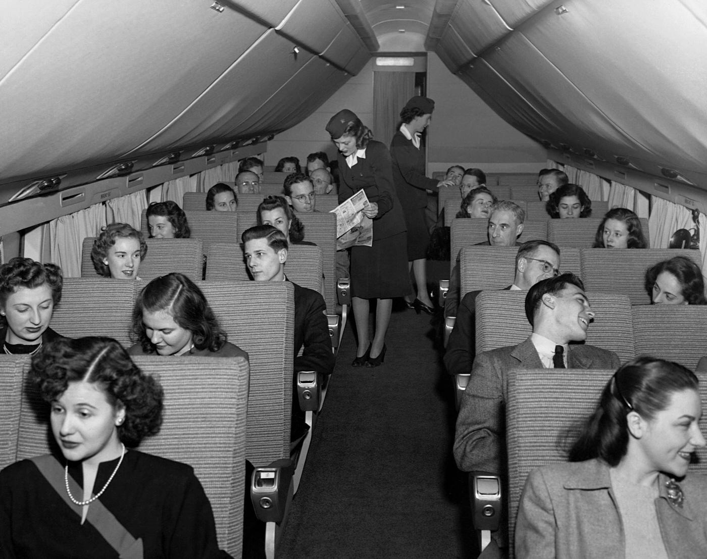 The interior of a Douglas DC-6 passenger airliner is shown, featuring passengers and flight attendants on a flight.