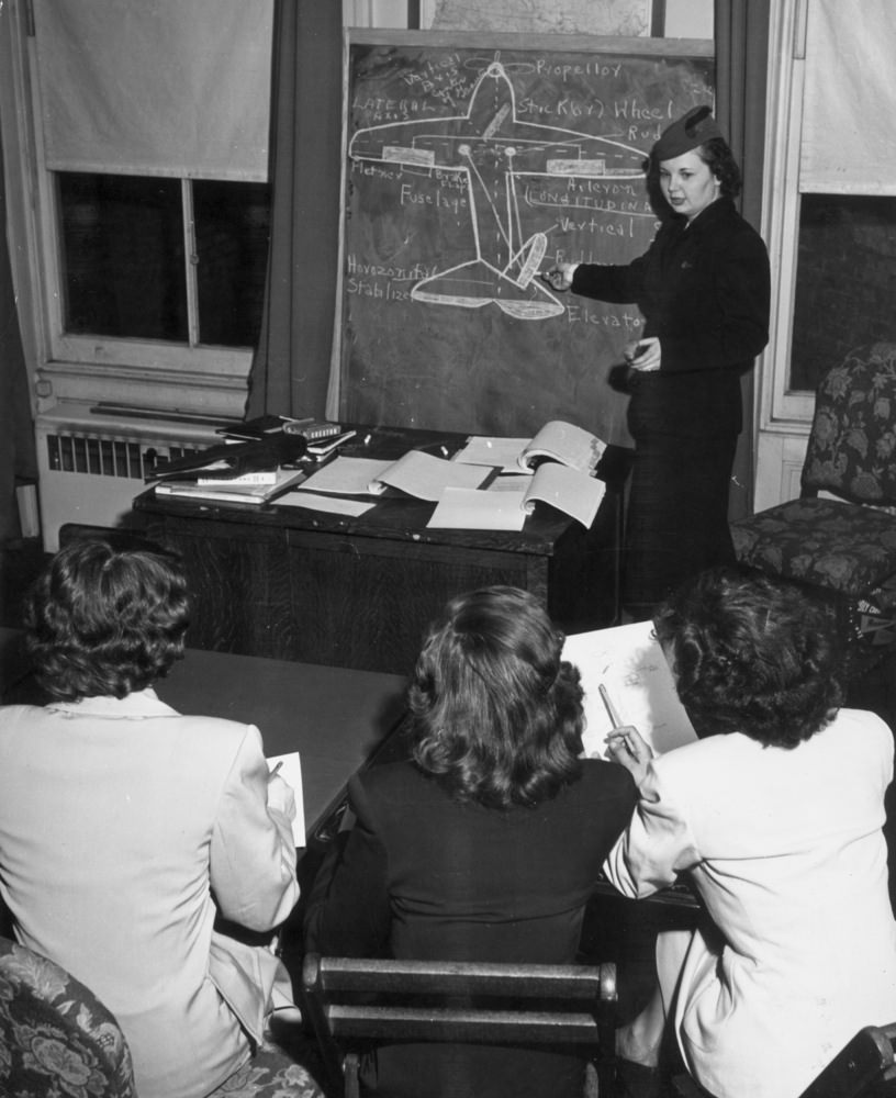 A female instructor stands at a chalkboard, pointing to a chalk diagram of an airplane while female students take notes at their desks during a stewardess training school, 1950.
