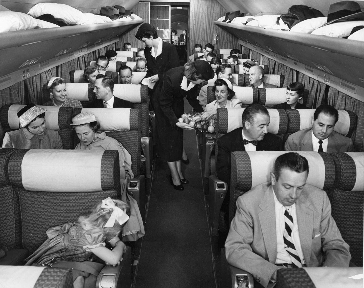 In 1955, an interior view of the passenger cabin of a Douglas DC-7 Mainliner airplane shows a pair of flight attendants serving travelers.