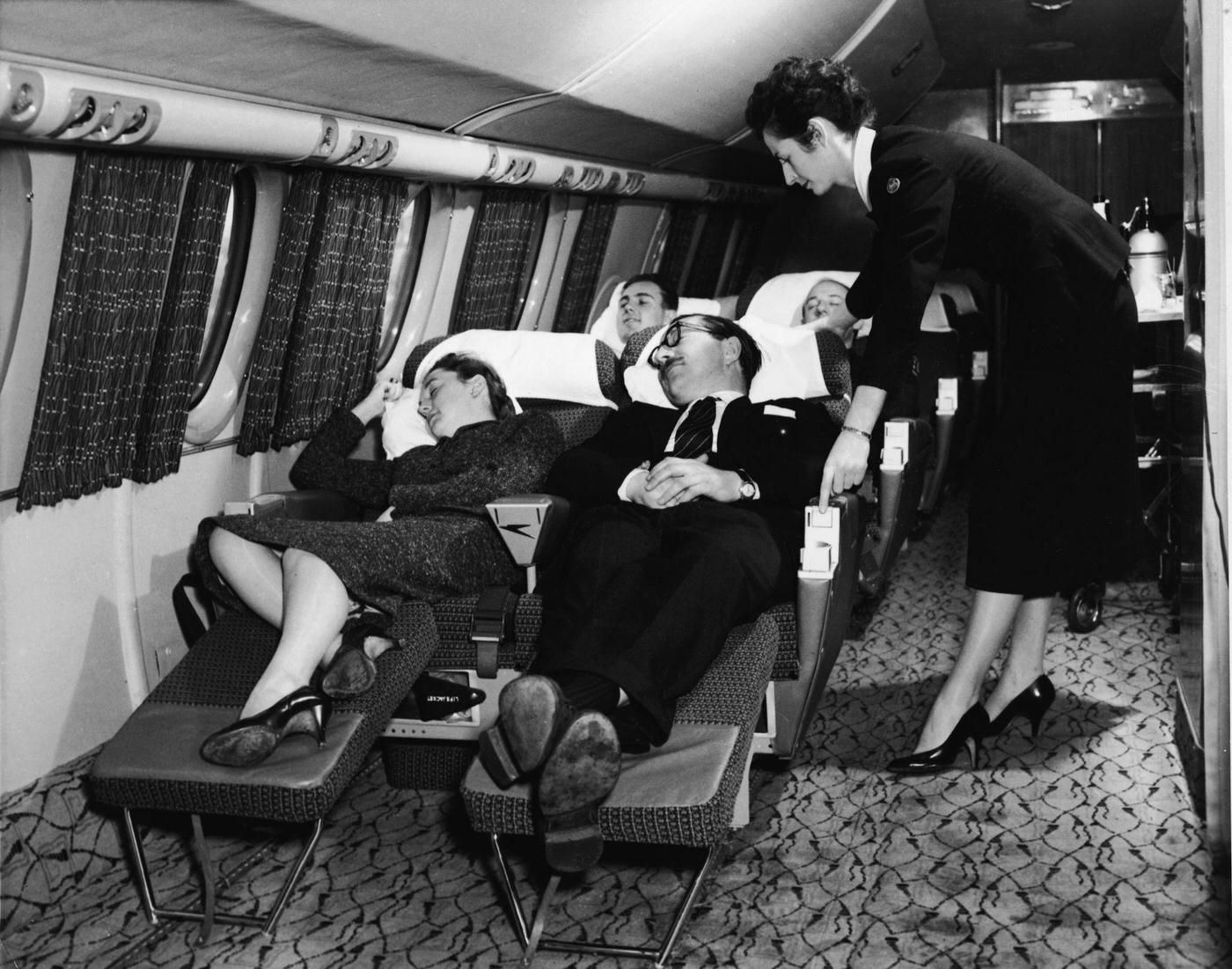 In the 1950s, an interior view of the first-class compartment of a commercial passenger plane shows a flight attendant bending forward to adjust the seat of a sleeping man.
