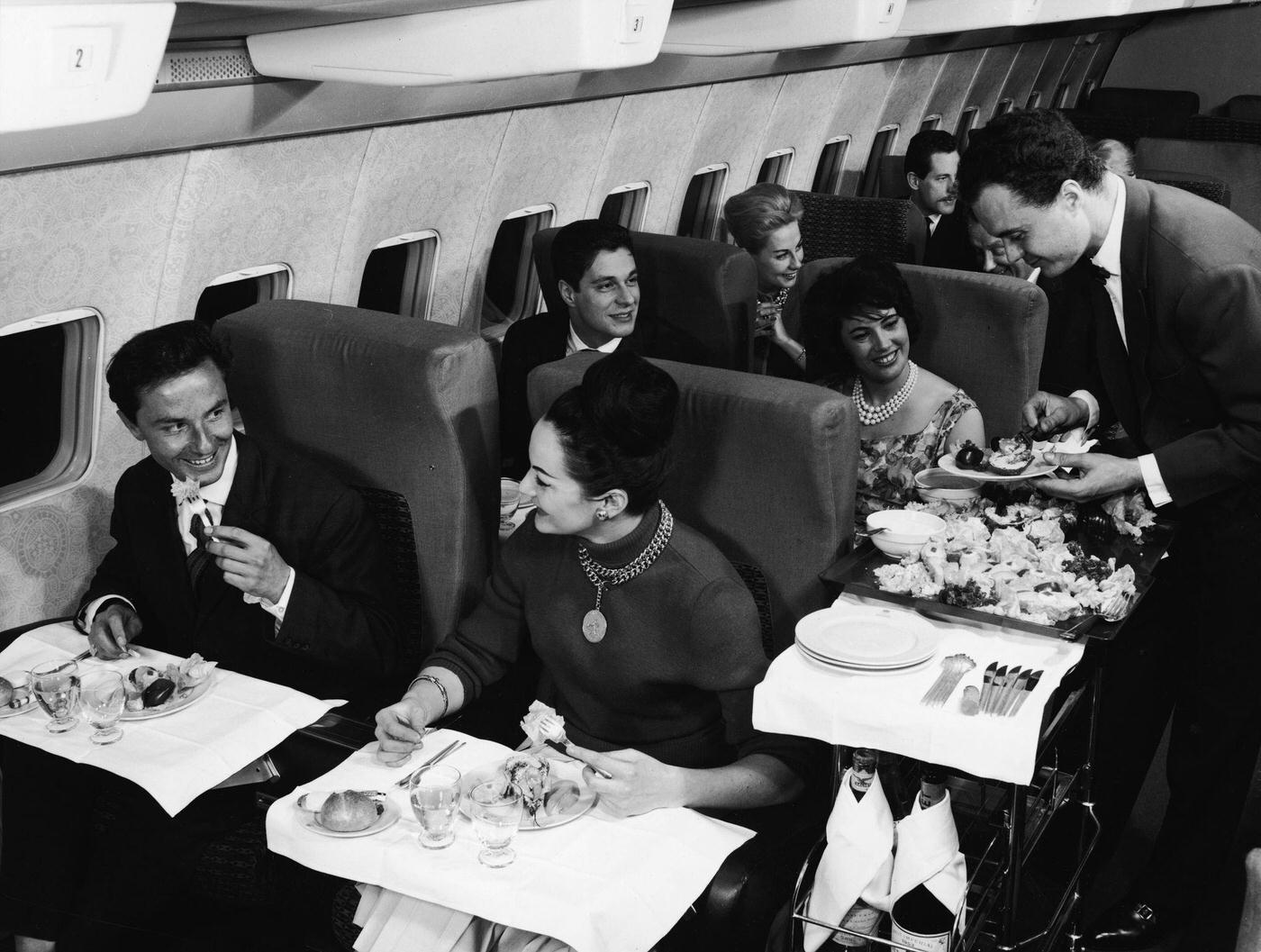 In the 1950s, a first-class compartment of a commercial passenger plane shows a well-dressed couple in the foreground smiling and enjoying their meal, while behind them a flight attendant in a bow tie serves another happy couple.