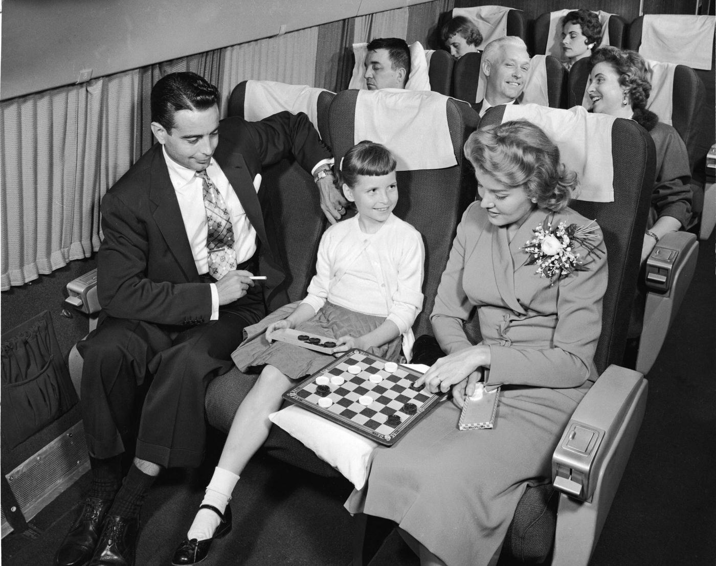 In the 1950s, an interior view of a commercial passenger plane shows a woman contemplating a move in a checkers game she is losing to a young girl, while a man with a cigarette watches them.