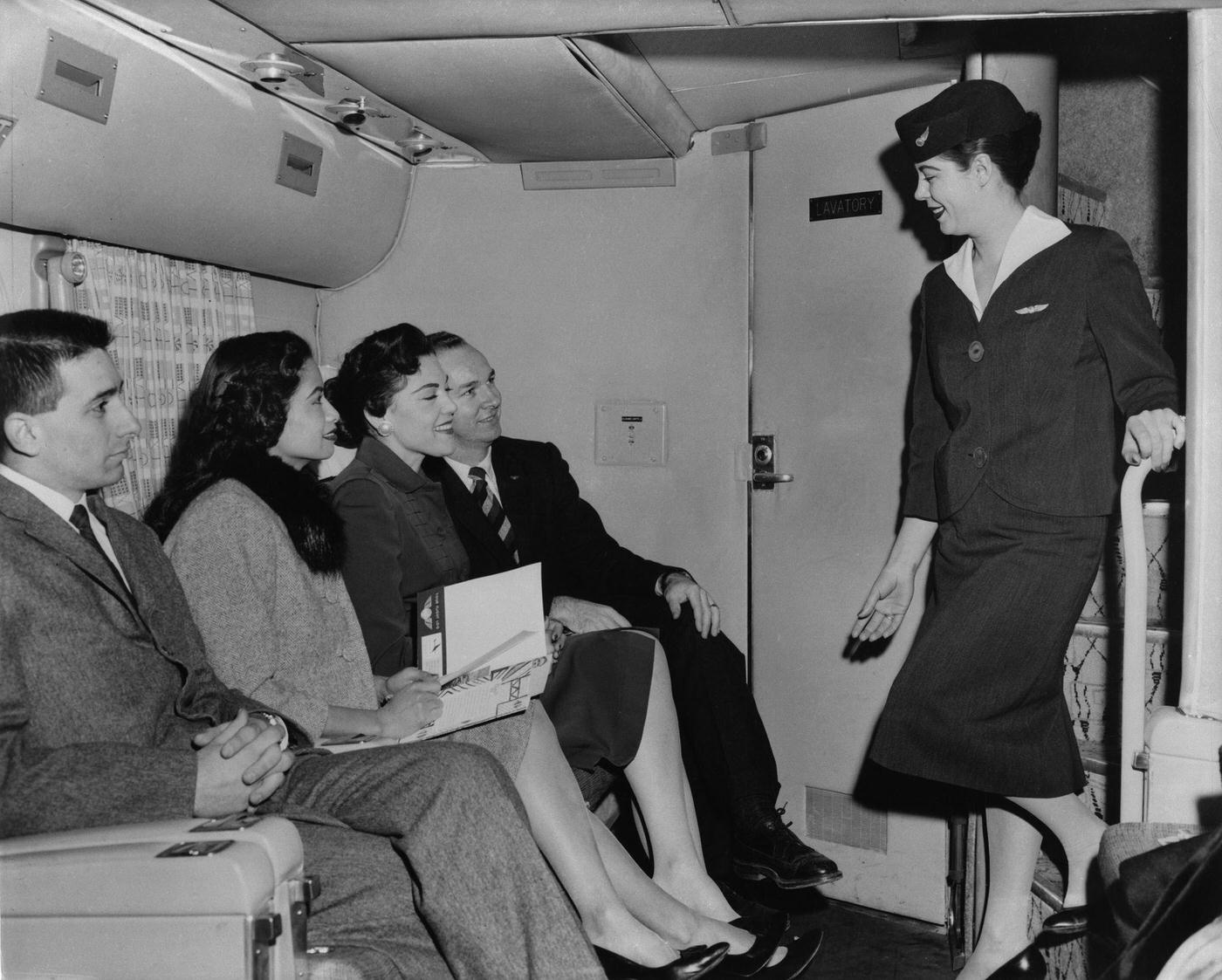 During a flight on a Transocean Air Lines Boeing 377 Stratocruiser in the mid 1950s, an air hostess takes time to chat with passengers and ensure their comfort during the flight.